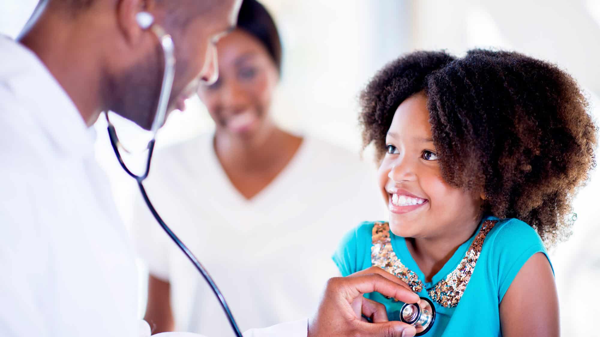 Pediatrician uses a stethoscope to listen to the heart of a smiling school-age child.